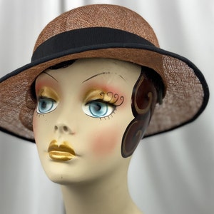 Pu&ograve; includere: Un cappello di paglia marrone con una fascia nera. Il cappello ha un'ampia tesa ed &egrave; posto su una testa di manichino.