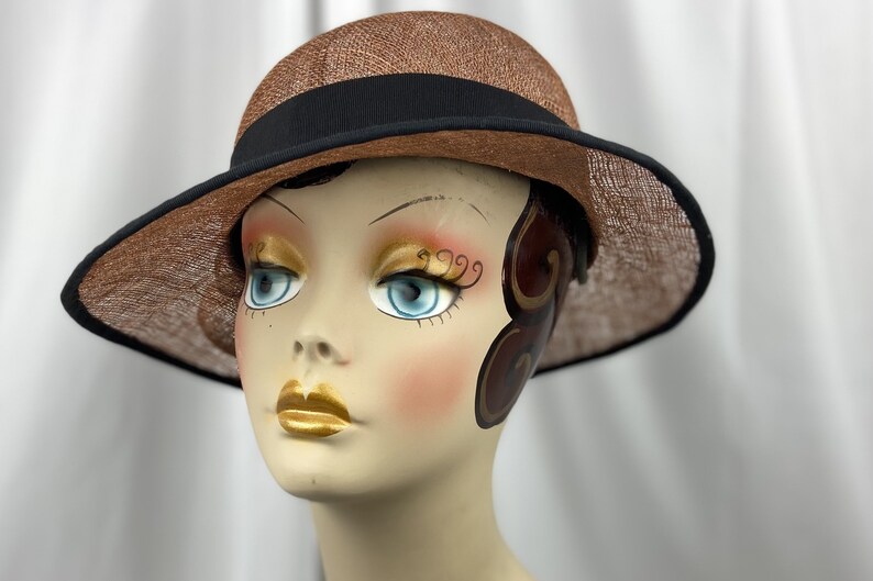 Pu&ograve; includere: Un cappello di paglia marrone con una fascia nera. Il cappello ha un'ampia tesa ed &egrave; posto su una testa di manichino.