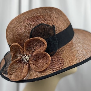 Pu&ograve; includere: Un cappello di paglia marrone e nero con un grande fiocco di fiori marroni. Il cappello ha un'ampia tesa e un nastro nero.