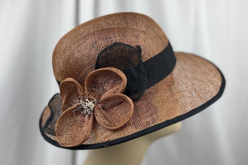 Pu&ograve; includere: Un cappello di paglia marrone e nero con un grande fiocco di fiori marroni. Il cappello ha un'ampia tesa e un nastro nero.