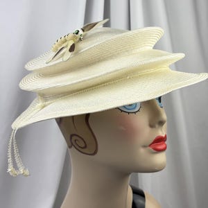 May include: A cream-colored tiered hat with a structured, multi-layered brim design. The hat features a decorative dragonfly ornament on the top, characterized by cream and brown wings with green accents, and includes thin dangling beaded tassels.