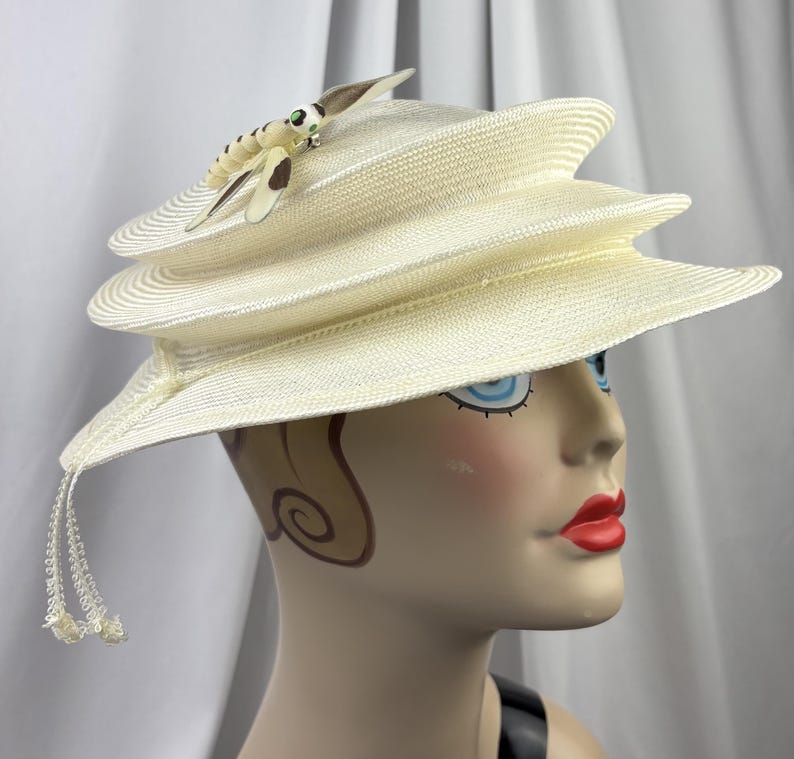 May include: A cream-colored tiered hat with a structured, multi-layered brim design. The hat features a decorative dragonfly ornament on the top, characterized by cream and brown wings with green accents, and includes thin dangling beaded tassels.
