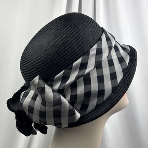 May include: A black cloche hat with a woven texture. The hat features a wide brim and is adorned with a black and white checkered fabric band and bow. The hat is displayed on a mannequin head.