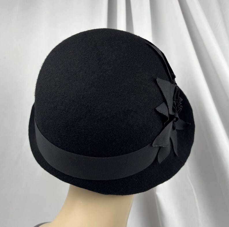 May include: A solid black felt cloche hat featuring a wide grosgrain ribbon band encircling the base. The hat is adorned with a decorative cluster of black fabric petals and leaves attached to the side, creating a textured floral accent.