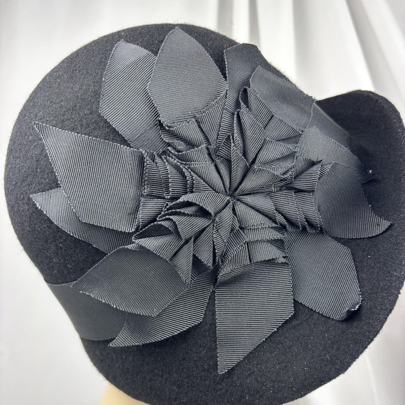 May include: A black felt hat featuring a large, decorative flower ornament made from layered black grosgrain ribbon. The ribbon petals are arranged in a dense, multi-layered pattern that covers the crown of the hat, creating a textured appearance.