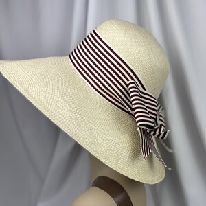 Pu&ograve; includere: Cappello di paglia a tesa larga di colore crema. Il cappello presenta un nastro a righe bianche e bordeaux, legato a fiocco. Il cappello &egrave; esposto su una testa di manichino.