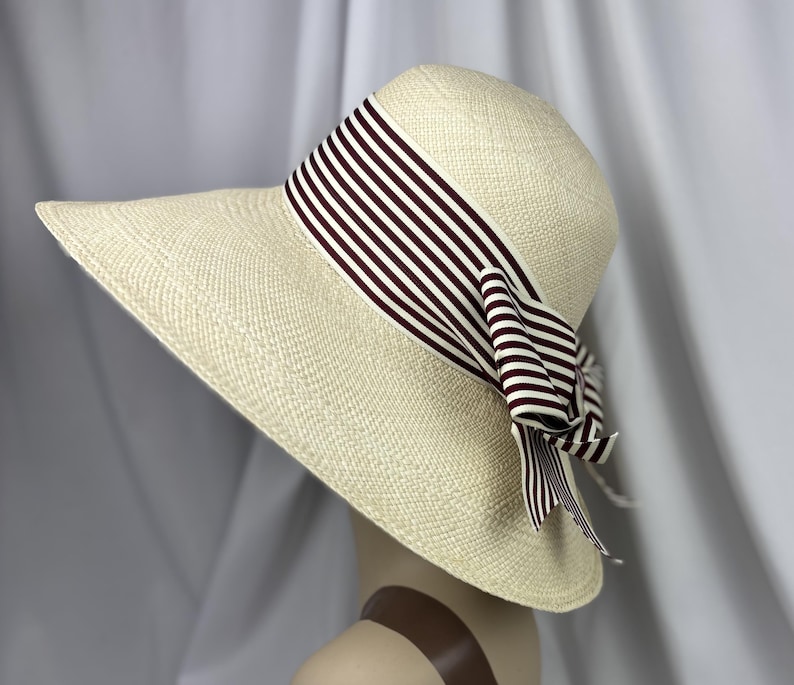 Pu&ograve; includere: Cappello di paglia a tesa larga di colore crema. Il cappello presenta un nastro a righe bianche e bordeaux, legato a fiocco. Il cappello &egrave; esposto su una testa di manichino.