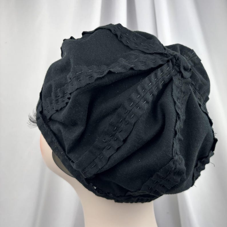May include: A black beret-style hat featuring a series of radiating, stitched fabric panels. The design includes decorative, raw-edged fabric strips sewn onto the surface in a starburst pattern, creating a textured, gathered appearance.
