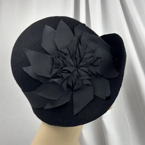 May include: A black felt cloche hat featuring a large, decorative flower made from folded black grosgrain ribbon. The hat has a smooth, rounded crown and a small, slightly upturned brim, with the ribbon flower centered on the side.
