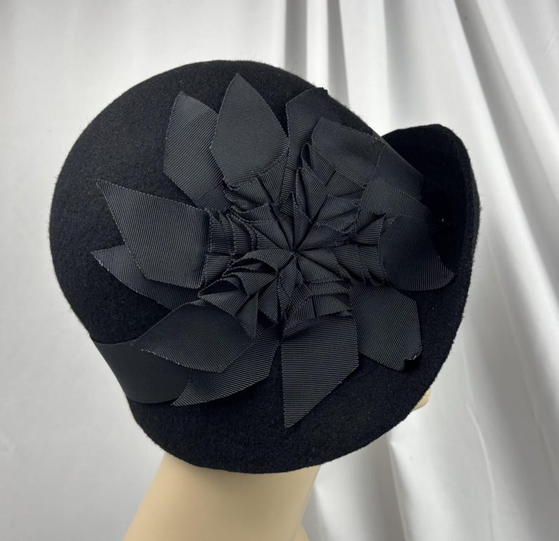 May include: A black felt cloche hat featuring a large, decorative flower made from folded black grosgrain ribbon. The hat has a smooth, rounded crown and a small, slightly upturned brim, with the ribbon flower centered on the side.