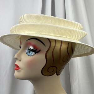 May include: A cream-colored straw hat featuring a multi-tiered, layered crown design and a wide, slightly downturned brim. The hat is constructed from a finely woven material with a consistent texture throughout the layered structure.