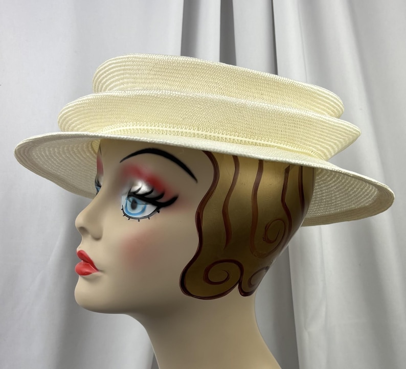 May include: A cream-colored straw hat featuring a multi-tiered, layered crown design and a wide, slightly downturned brim. The hat is constructed from a finely woven material with a consistent texture throughout the layered structure.