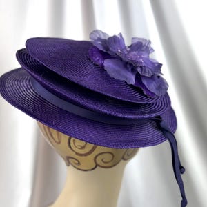 May include: A layered purple hat with a decorative flower. The hat features a ribbon band and a long, flowing ribbon. The hat is displayed on a mannequin head. The hat is a deep shade of purple, suitable for special occasions.