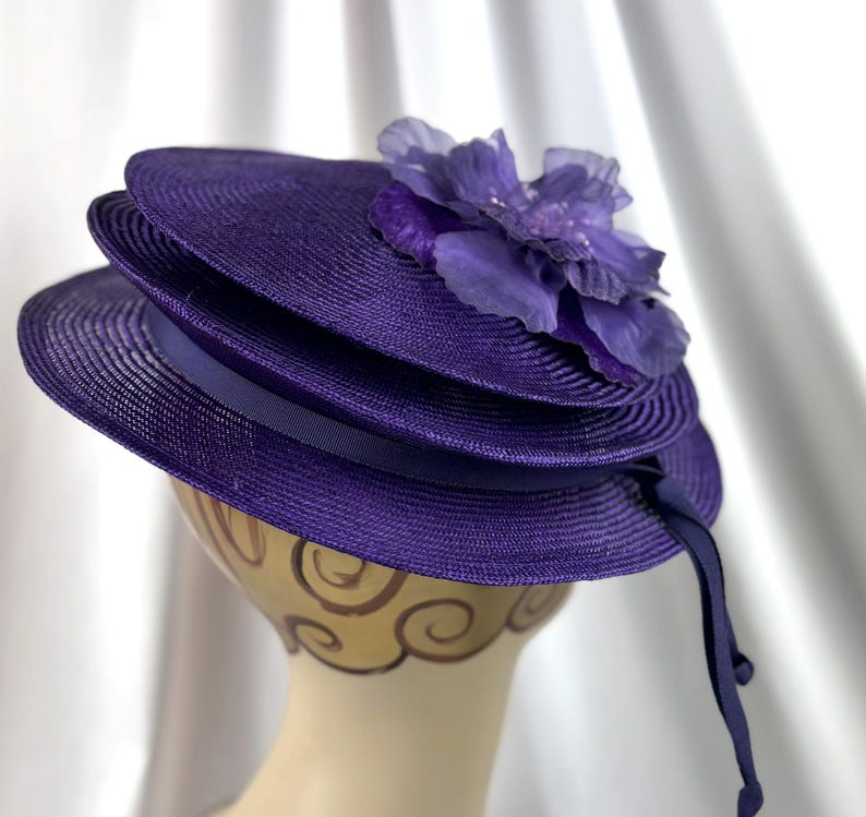 May include: A layered purple hat with a decorative flower. The hat features a ribbon band and a long, flowing ribbon. The hat is displayed on a mannequin head. The hat is a deep shade of purple, suitable for special occasions.