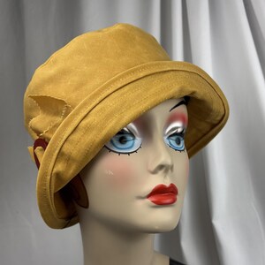 Puede incluir: Un sombrero de pescador color amarillo mostaza con ala ancha inclinada hacia abajo y acabado de tela texturizada. El sombrero presenta un detalle decorativo lateral que consiste en un peque&ntilde;o lazo de tela y un anillo circular rojo oscuro.