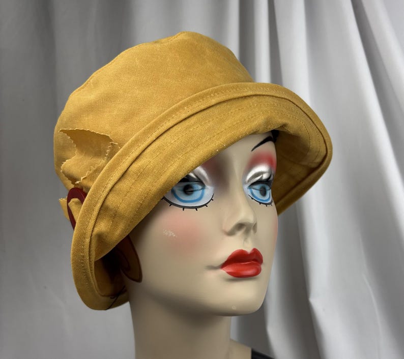 Puede incluir: Un sombrero de pescador color amarillo mostaza con ala ancha inclinada hacia abajo y acabado de tela texturizada. El sombrero presenta un detalle decorativo lateral que consiste en un peque&ntilde;o lazo de tela y un anillo circular rojo oscuro.