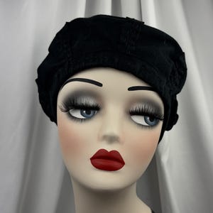 May include: A solid black beret featuring a structured, multi-paneled crown design with visible stitching details along the seams. The hat is crafted from a dark fabric and sits securely on the head, showcasing a classic rounded silhouette.