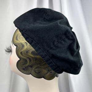 May include: A black beret featuring visible tonal stitching details along the crown and the brim. The hat is displayed on a mannequin head with stylized dark hair and long eyelashes, showcasing the soft fabric texture and rounded shape.