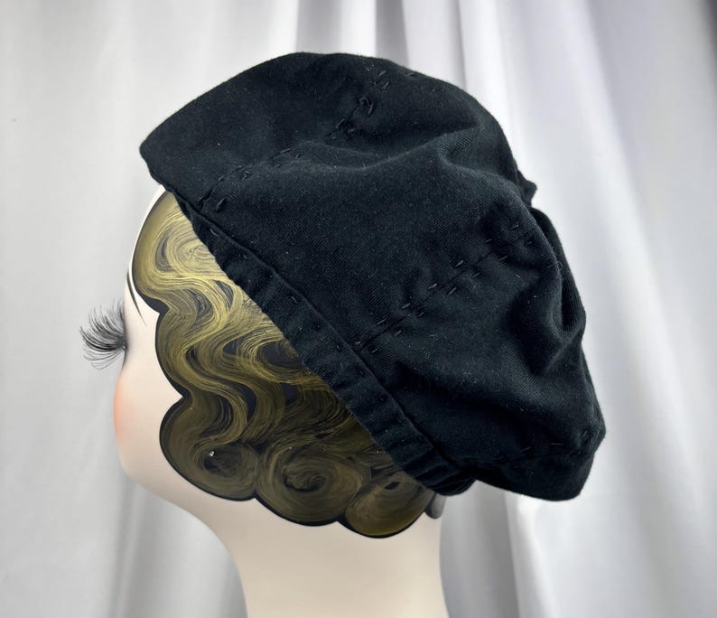 May include: A black beret featuring visible tonal stitching details along the crown and the brim. The hat is displayed on a mannequin head with stylized dark hair and long eyelashes, showcasing the soft fabric texture and rounded shape.