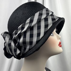 May include: A black cloche hat with a wide brim and a black and white chequered band. The hat is made of woven material and features a decorative bow on the side. The hat is displayed on a mannequin head.