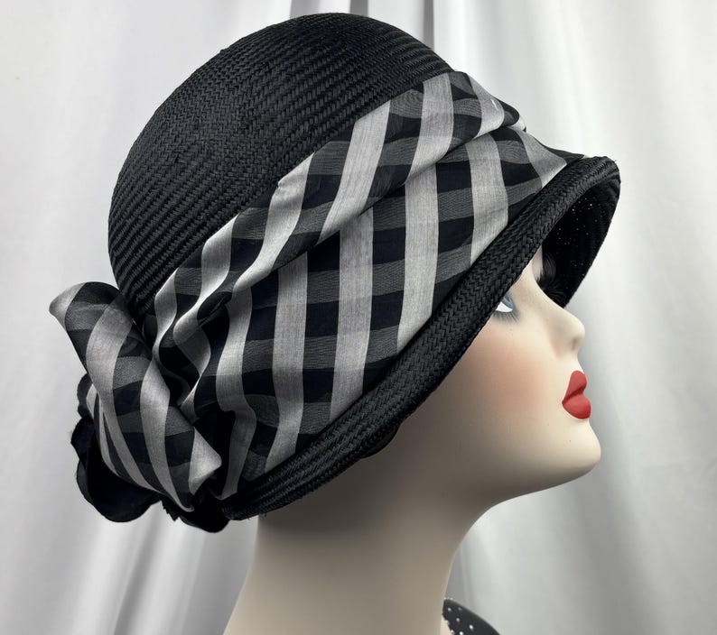 May include: A black cloche hat with a wide brim and a black and white chequered band. The hat is made of woven material and features a decorative bow on the side. The hat is displayed on a mannequin head.