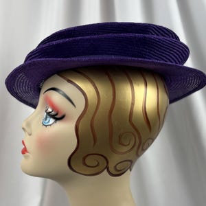 May include: A vintage purple hat with a structured design. The hat has a wide brim and a layered crown. The hat is displayed on a mannequin head with painted features and gold-coloured hair.