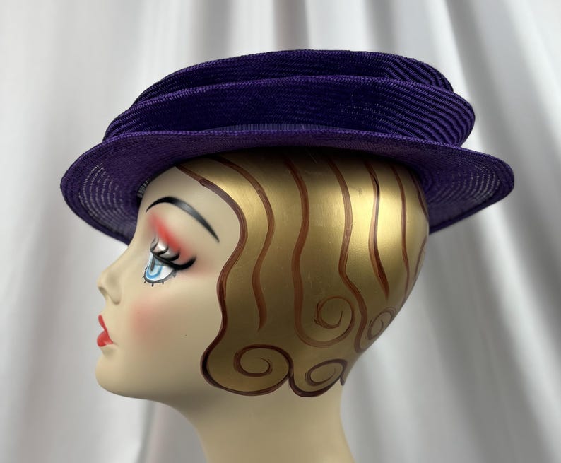 May include: A vintage purple hat with a structured design. The hat has a wide brim and a layered crown. The hat is displayed on a mannequin head with painted features and gold-coloured hair.