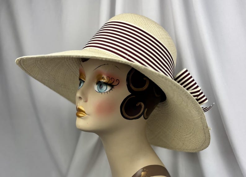 Pu&ograve; includere: Cappello di paglia a tesa larga con corona e tesa color crema. Il cappello presenta un nastro a righe bordeaux scuro e bianco, con un fiocco abbinato sul retro. Il cappello &egrave; esposto su una testa di manichino.