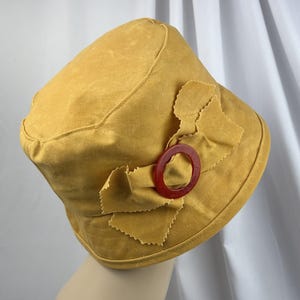 Puede incluir: Un sombrero de pescador color amarillo mostaza con un detalle de lazo de tela en el lateral. El lazo est&aacute; sujeto con una anilla circular de pl&aacute;stico rojo oscuro. El sombrero est&aacute; confeccionado en tela mate de color s&oacute;lido con costuras visibles.