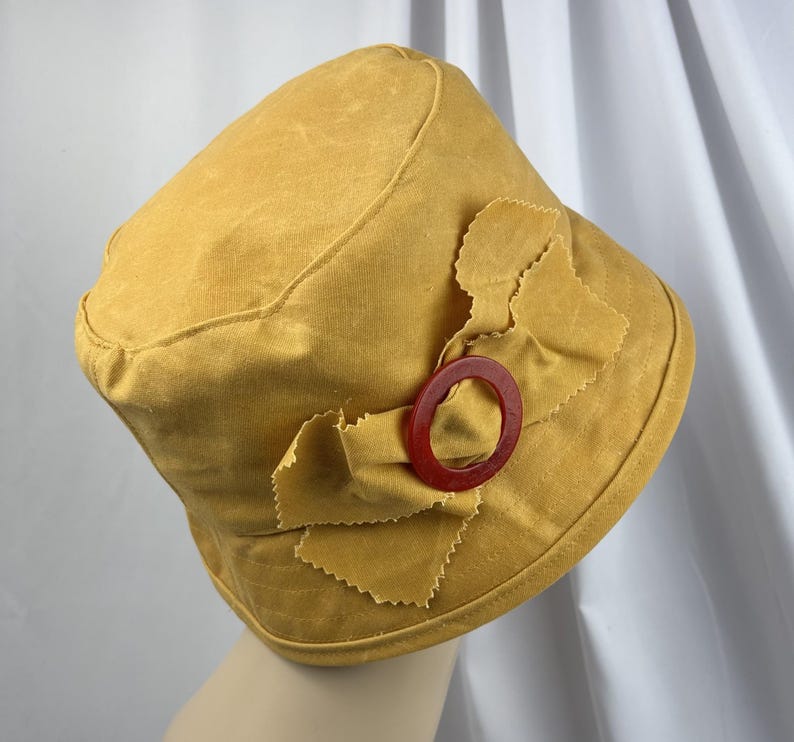 Puede incluir: Un sombrero de pescador color amarillo mostaza con un detalle de lazo de tela en el lateral. El lazo est&aacute; sujeto con una anilla circular de pl&aacute;stico rojo oscuro. El sombrero est&aacute; confeccionado en tela mate de color s&oacute;lido con costuras visibles.