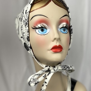 May include: A white headscarf with a black floral pattern and a checkered border. The scarf is tied under the chin with matching ties. The head covering is displayed on a mannequin head with blue eyes and red lips.