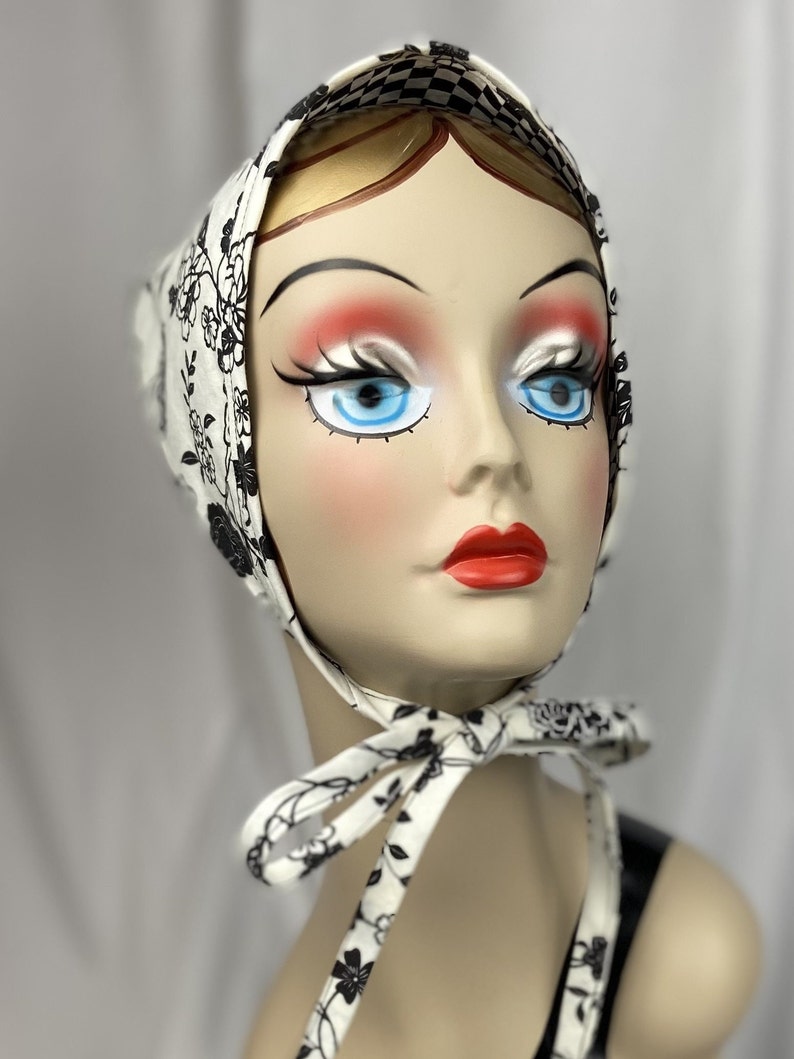 May include: A white headscarf with a black floral pattern and a checkered border. The scarf is tied under the chin with matching ties. The head covering is displayed on a mannequin head with blue eyes and red lips.