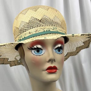 May include: A straw hat with a wide, scalloped brim and a woven crown. The hat features a teal ribbon and decorative accents. The hat is displayed on a mannequin head with blue eyes and red lipstick.