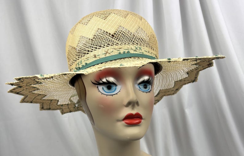 May include: A straw hat with a wide, scalloped brim and a woven crown. The hat features a teal ribbon and decorative accents. The hat is displayed on a mannequin head with blue eyes and red lipstick.