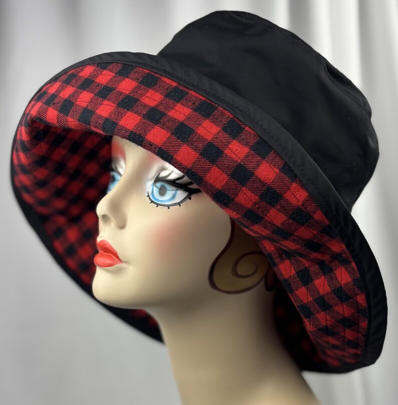 Pu&ograve; includere: Cappello da pescatore nero con fodera a quadri rossi e neri. Il cappello ha un'ampia tesa ed &egrave; esposto su una testa di manichino. &Egrave; un accessorio elegante.