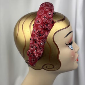 May include: A red fabric headband with a paisley pattern and small silver beads. The headband is wrapped around a mannequin head with gold paint and a vintage-style face.