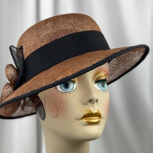 Pu&ograve; includere: Un cappello di paglia marrone con un nastro nero e un bordo nero. Il cappello ha un'ampia tesa e una piccola corona.