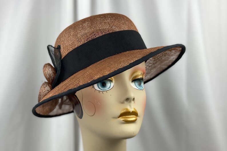 Pu&ograve; includere: Un cappello di paglia marrone con un nastro nero e un bordo nero. Il cappello ha un'ampia tesa e una piccola corona.