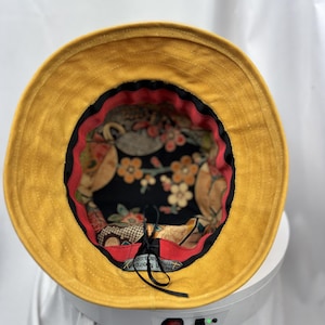 Puede incluir: Un sombrero de pescador color amarillo mostaza con ala ancha, que presenta un forro interior negro decorado con un estampado floral en tonos naranja, blanco y verde. Una banda de tela roja bordea el borde interior y tiene una etiqueta negra.
