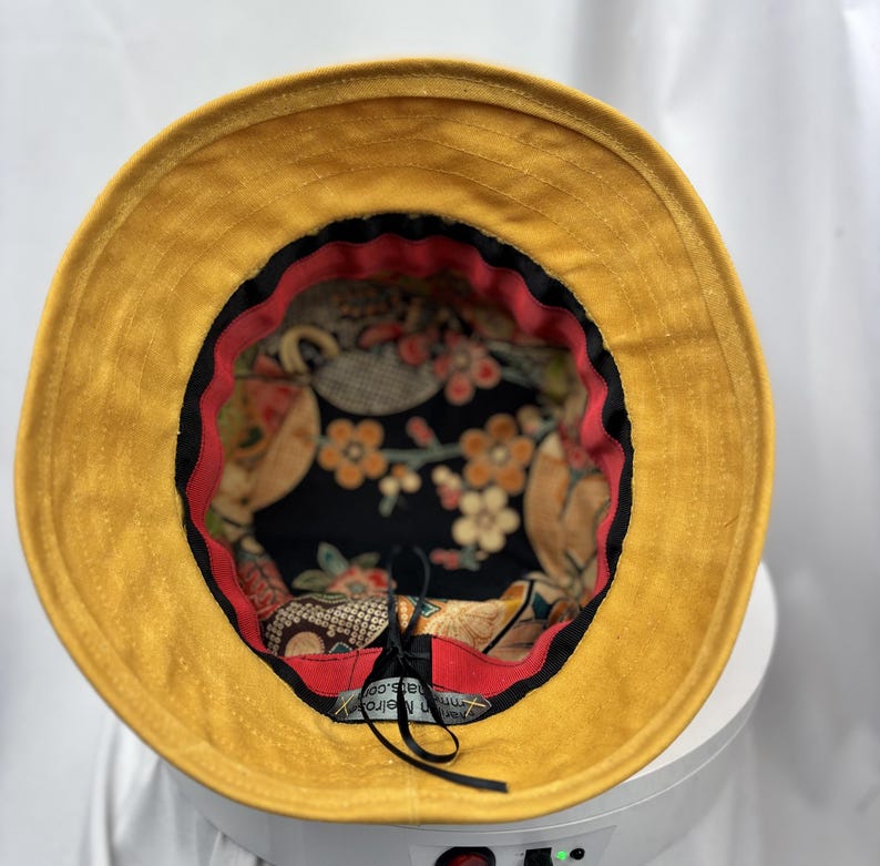 Puede incluir: Un sombrero de pescador color amarillo mostaza con ala ancha, que presenta un forro interior negro decorado con un estampado floral en tonos naranja, blanco y verde. Una banda de tela roja bordea el borde interior y tiene una etiqueta negra.