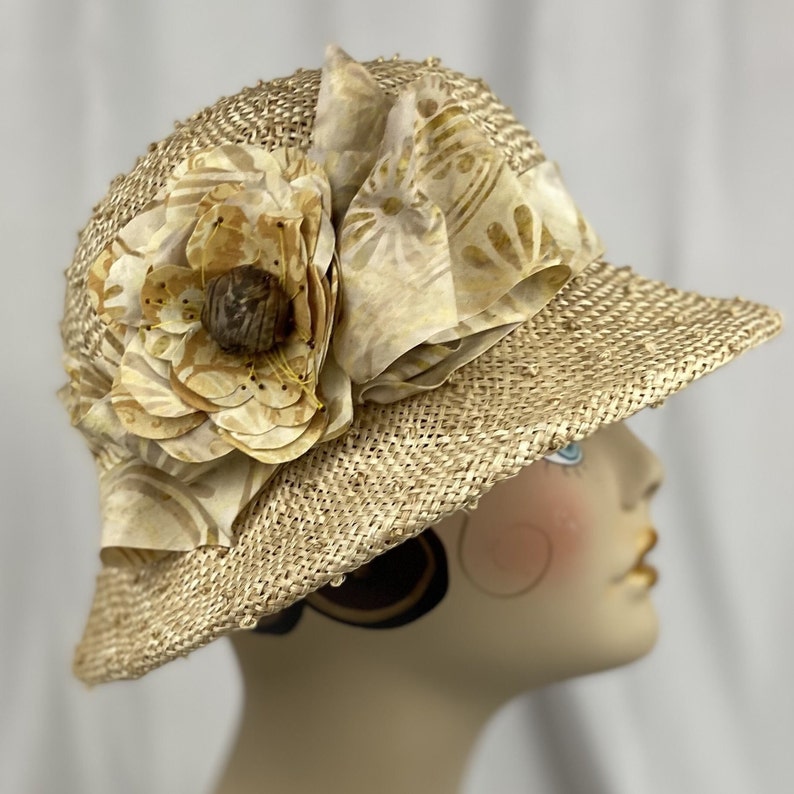Puede incluir: Un sombrero de paja de ala ancha con una gran flor beige como adorno. La flor tiene un centro marr&oacute;n y est&aacute; hecha de tela con un patr&oacute;n sutil.