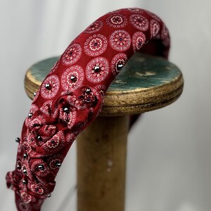 May include: A burgundy headband with a repeating circular pattern in white and navy blue. The headband is embellished with small, silver-colored studs along the edge. The accessory is resting on a wooden spool.