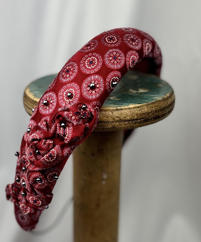 May include: A burgundy headband with a repeating circular pattern in white and navy blue. The headband is embellished with small, silver-colored studs along the edge. The accessory is resting on a wooden spool.