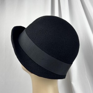 May include: A black felt cloche hat featuring a rounded crown and a narrow, slightly upturned brim. A wide, smooth black grosgrain ribbon band encircles the base of the crown, providing a subtle contrast in texture against the matte felt material.