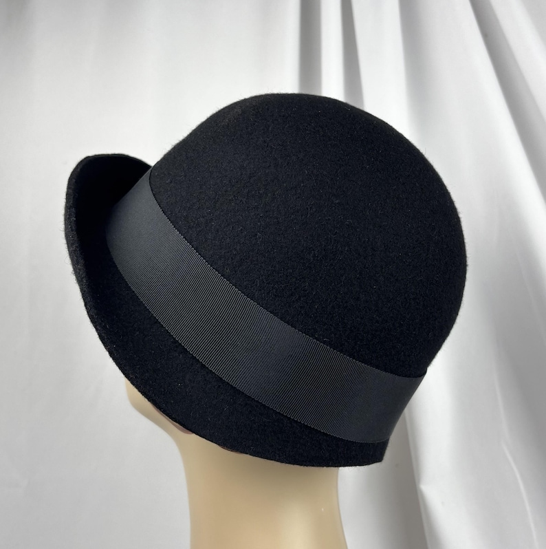 May include: A black felt cloche hat featuring a rounded crown and a narrow, slightly upturned brim. A wide, smooth black grosgrain ribbon band encircles the base of the crown, providing a subtle contrast in texture against the matte felt material.