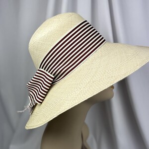 Pu&ograve; includere: Un cappello di paglia a tesa larga con una trama color crema. Il cappello presenta un nastro a righe bordeaux scuro e bianco legato a fiocco. Il cappello &egrave; esposto su una testa di manichino.
