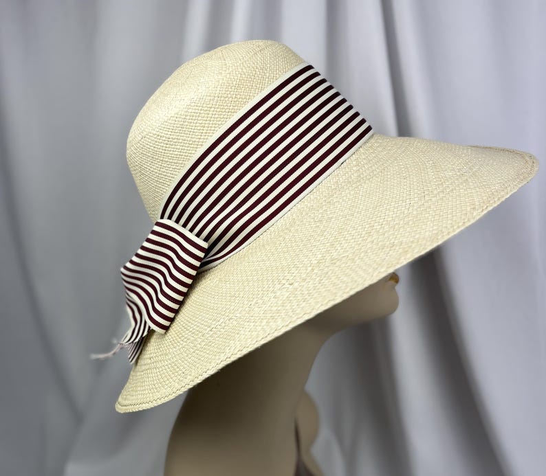 Pu&ograve; includere: Un cappello di paglia a tesa larga con una trama color crema. Il cappello presenta un nastro a righe bordeaux scuro e bianco legato a fiocco. Il cappello &egrave; esposto su una testa di manichino.