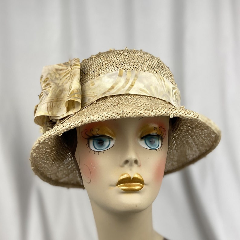 Puede incluir: Un sombrero de paja de ala ancha con una cinta de color crema con un patr&oacute;n floral. El sombrero est&aacute; sentado en la cabeza de un maniqu&iacute;.