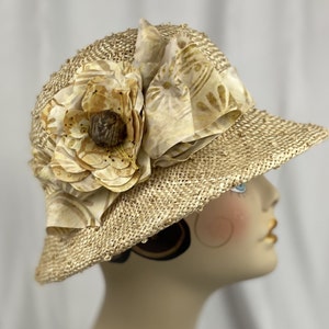 Puede incluir: Un sombrero de paja de ala ancha con una gran flor como adorno. La flor est&aacute; hecha de tela y tiene una combinaci&oacute;n de colores marr&oacute;n claro y beige.
