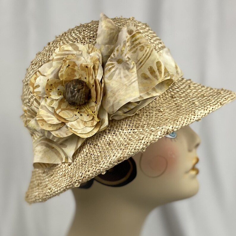 Puede incluir: Un sombrero de paja de ala ancha con una gran flor como adorno. La flor est&aacute; hecha de tela y tiene una combinaci&oacute;n de colores marr&oacute;n claro y beige.